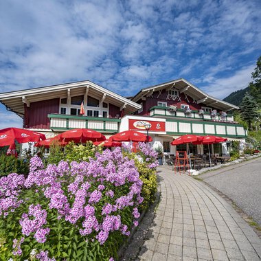 Ein malerisches alpenländisches Haus mit roten Sonnenschirmen und bunten Blumen. Der Himmel ist blau mit einigen Wolken. | © Gasthof Alpenblick | Theodor Pinn