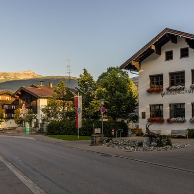 Eine malerische Straßenansicht eines kleinen Dorfes in den Alpen, umgeben von Bergen. Die charmanten Häuser sind mit Blumen geschmückt und schaffen eine einladende Atmosphäre. | © Kleinwalsertal Tourismus |  Steffen Berschin