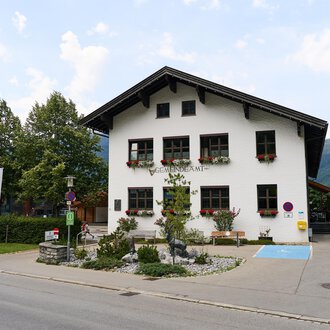 Ein traditionelles, zweigeschossiges Haus mit dekorativen Fenstern und einem klaren Himmel. Umgeben von Bäumen und einer ruhigen Straße. | © Kleinwalsertal Tourismus | Oliver Farys