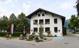 Ein traditionelles, zweigeschossiges Haus mit dekorativen Fenstern und einem klaren Himmel. Umgeben von Bäumen und einer ruhigen Straße. | © Kleinwalsertal Tourismus | Oliver Farys