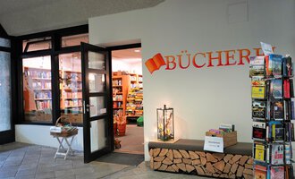 An inviting entrance to a library with a large selection of books inside. Next to it stands a bookshelf and a small seating area. | © Kleinwalsertal Tourismus | S. Guggenberger
