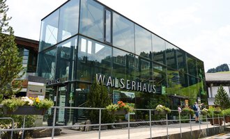 A modern building with a glass facade stands in the foreground. Surrounded by trees and flowers, it conveys an inviting impression. | © Kleinwalsertal Tourismus | Oliver Farys