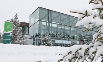 A modern building with large glass windows in the snow. Surrounded by snow-covered trees, it showcases a winter atmosphere. | © Kleinwalsertal Tourismus | Oliver Farys