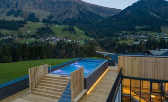A modern building with a wooden deck and a pool that is embedded in the landscape. In the background, the mountains and the surrounding nature are visible.