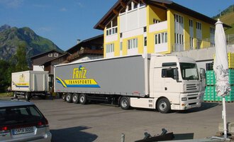 Ein Lkw von Fritz Transporte steht auf einem Parkplatz vor einem großen Gebäude. Im Hintergrund sind grüne Berge zu sehen. | © Getränke Service Fritz | Helmut Fritz