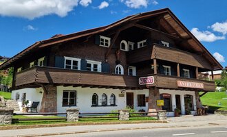 Ein charmantes, traditionelles Chalet aus Holz mit mehreren Balkonen und Fenstern. Der Himmel ist blau und voller Wolken, während der grüne Rasen das Gebäude umgibt. | © Antik und Kunst im Malerhäusl Herbert Stumvoll