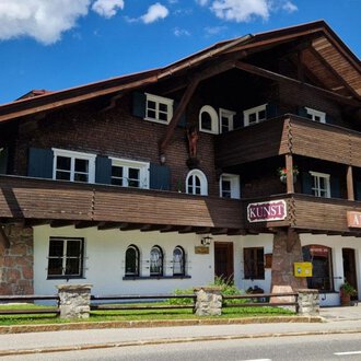 A charming, traditional wooden chalet with multiple balconies and windows. The sky is blue and filled with clouds, while the green lawn surrounds the building. | © Antik und Kunst im Malerhäusl Herbert Stumvoll