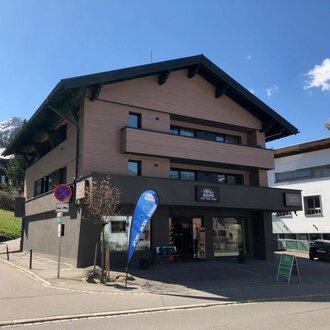 A modern building with several floors is located on a quiet street. In the background, mountains and additional buildings can be seen. | © Sport Hilbrand GmbH