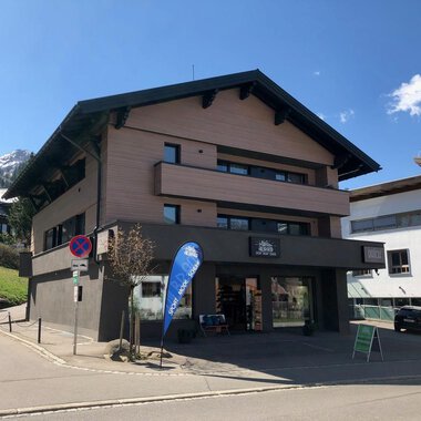 Ein modernes Gebäude mit mehreren Etagen liegt an einer ruhigen Straße. Im Hintergrund sind Berge und weitere Gebäude zu sehen. | © Sport Hilbrand GmbH