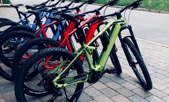 Mehrere Mountainbikes in unterschiedlichen Farben stehen auf einem Kiesweg. Im Hintergrund ist ein Holzhaus und eine grüne Wiese zu sehen. | © Sport Hilbrand GmbH