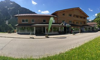 A modern wooden building in the mountains with a clear blue sky. In front of the building, a green meadow stretches out. | © Sport Hilbrand GmbH