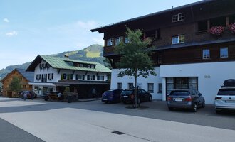 A quiet village with traditional houses and cars on the street. Green hills are visible in the background. | © Kleinwalsertal Tourismus | Nicole Lughammer