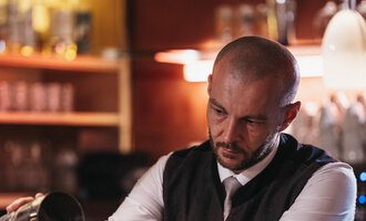 Ein Barkeeper schenkt einen Drink in ein Glas ein. Im Hintergrund sind Gläser und eine stilvolle Bar zu sehen. | © Naturhotel Chesa Valisa | Heldentheater