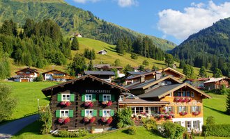 Eine malerische Landschaft mit traditionellen Häusern in den Bergen. Üppige grüne Wiesen und Wälder umgeben die Gebäude. | © Alpenhof Jäger | Werbewind