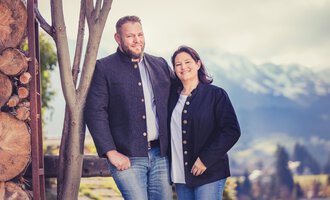 Ein Paar steht lächelnd vor einer malerischen Berglandschaft. Beide tragen traditionelle Kleidung und wirken freundlich und entspannt. | © Alpenhof Jäger | Werbewind
