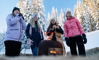 Eine Gruppe von vier Frauen steht im Schnee und genießt die winterliche Atmosphäre. Sie lächeln, während sie sich um ein kleines Feuer versammeln. | © Kleinwalsertal Tourismus | Oliver Farys