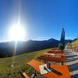 Eine schöne Berglandschaft mit Sonne, die über den Gipfeln scheint. Im Vordergrund stehen Holzsitze und Tische mit grünen Sonnenschirmen. | © Ifenhütte | Petra Diener