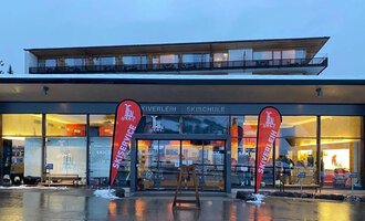 Ein modernes Skizentrum mit großen Fenstern und rot-weißen Fahnen. Der Eingangsbereich ist einladend und spiegeln die winterliche Atmosphäre wider. | © Ifensport | Thomas MARTIN