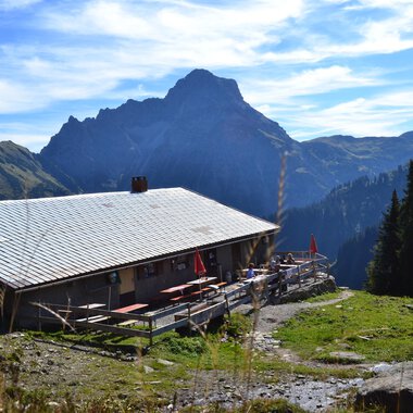 An alpine cabin surrounded by mountains and forests. The sky is clear with a few clouds. | © Kleinwalsertal Tourismus