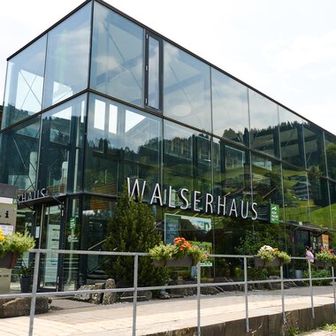 Ein modernes Gebäude namens Walserhaus in einer bergigen Umgebung. Umgeben von Bäumen und unter einem klaren Himmel. | © Kleinwalsertal Tourismus | Oliver Farys