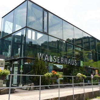 Ein modernes Gebäude namens Walserhaus in einer bergigen Umgebung. Umgeben von Bäumen und unter einem klaren Himmel. | © Kleinwalsertal Tourismus | Oliver Farys