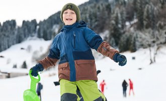 A cheerful boy is wearing a colorful winter jacket and holding a sled in his hand. In the background, you can see snow-covered landscapes and other people in the snow. | © Kleinwalsertal Tourismus | Oliver Farys