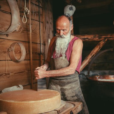 Ein älterer Mann mit langen weißen Haaren und Bart arbeitet in einer Holzwerkstatt. Im Hintergrund ist ein großer Kessel zu sehen. | © Käsestadl Kleinwalsertal | Nadine Paul