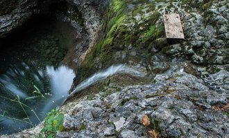 Eine felsige Klippe mit fließendem Wasser, das in einen tiefen, dunklen Pool mündet. Grüne Moose und Pflanzen wachsen zwischen den Steinen. | © Kleinwalsertal Tourismus | Andre Tappe
