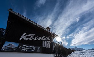 A clearly visible sign with the name "Kessler" on a foreground building. In the background, the sky and the mountains are visible. | © Sport Kessler GmbH