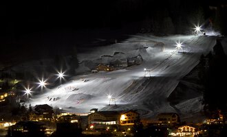 A lit ski slope at night with several skiers. In the foreground, cozy huts and a tranquil atmosphere can be seen. | © Sport Kessler GmbH