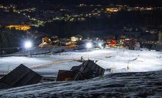 A illuminated ski slope at night with snow-covered slopes. In the background, the lights of a village are visible. | © Sport Kessler GmbH
