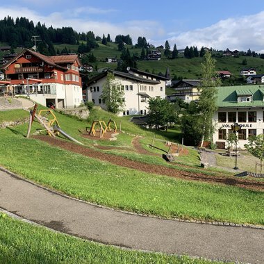 Eine malerische Landschaft mit grünen Wiesen und Spielplätzen. Im Hintergrund sind einige chaletartige Gebäude und Berge zu sehen. | © Kleinwalsertal Tourismus