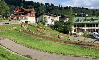 Eine malerische Landschaft mit grünen Wiesen und Spielplätzen. Im Hintergrund sind einige chaletartige Gebäude und Berge zu sehen. | © Kleinwalsertal Tourismus