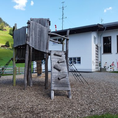 Ein Spielplatz mit einer Holzkletterstruktur und einer Rutsche. Im Hintergrund sieht man ein Haus und grüne Hügel. | © Kleinwalsertal Tourismus | N. Lughammer