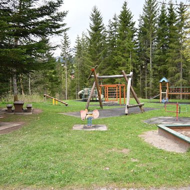 Ein Spielplatz in einem Wald mit mehreren Spielgeräten und einer Wiese. Im Hintergrund stehen hohe Bäume und eine Picknickbank. | © Gemeinde Mittelberg
