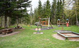 Ein Spielplatz in einem Wald mit mehreren Spielgeräten und einer Wiese. Im Hintergrund stehen hohe Bäume und eine Picknickbank. | © Gemeinde Mittelberg