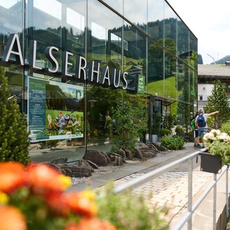 A modern building with a glass façade and the inscription "Walserhaus". In the foreground, colorful flowers are blooming, while in the background, the mountains and traditional buildings can be seen. | © Kleinwalsertal Tourismus | Oliver Farys