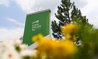 A sign with the inscription "Klein Walsertal" stands in the foreground. In the background, green trees and colorful flowers can be seen. | © Kleinwalsertal Tourismus | Oliver Farys