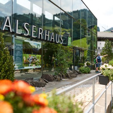 A modern building with a glass façade and the inscription "Walserhaus". In the foreground, colorful flowers are blooming, while in the background, the mountains and traditional buildings can be seen. | © Kleinwalsertal Tourismus | Oliver Farys