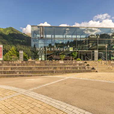 Ein modernes Gebäude mit Glasfassade und der Aufschrift "WALSERHAUS". Im Hintergrund sind grüne Berge und ein blauer Himmel sichtbar. | © Kleinwalsertal Tourismus | Steffen Berschin