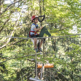 A boy is sitting on a rope in a high ropes course. The surroundings are surrounded by trees, and he is wearing a helmet and safety gear. | © OBERSTDORF · KLEINWALSERTAL BERGBAHNEN