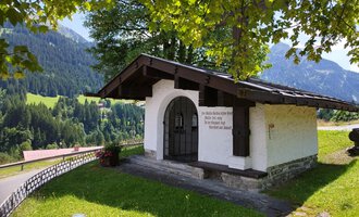 Ein kleines, weißes Gebäude mit einem giebelförmigen Dach steht in einer grünen Landschaft. Im Hintergrund sind sanfte Hügel und Bäume zu sehen. | © Kleinwalsertal Tourismus | Nicole Lughammer