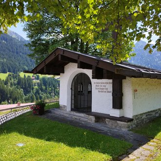 Ein kleines, weißes Gebäude mit einem giebelförmigen Dach steht in einer grünen Landschaft. Im Hintergrund sind sanfte Hügel und Bäume zu sehen. | © Kleinwalsertal Tourismus | Nicole Lughammer