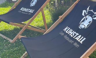 Zwei Liegestühle mit dem Logo „Kuhstall“ stehen auf einer Wiese. Im Hintergrund sind einige Gebäude und ein Willkommen-Schild sichtbar. | © Kuhstall | Uwe Nagel