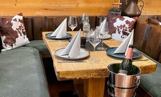 Ein gemütlicher Tisch in einem rustikalen Restaurant, dekoriert mit gefalteten Servietten und Gläsern. Eine Flasche Sekt steht in einem Kübel bereit. | © Kuhstall | Uwe Nagel