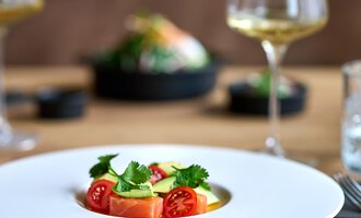 Ein stilvolles Gericht auf einem weißen Teller, dekoriert mit frischen Tomaten und Kräutern. Im Hintergrund sind Weingläser und eine weitere Speise zu sehen. | © La Vallée | Annette Sandner