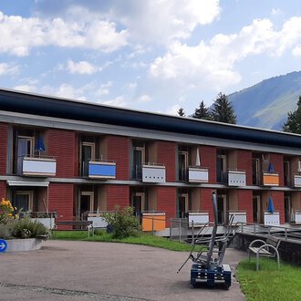Ein modernes Gebäude mit mehreren Etagen und Balkonen. Im Hintergrund sind grüne Hügel und ein blauer Himmel zu sehen. | © Kleinwalsertal Tourismus | Veronika Senn