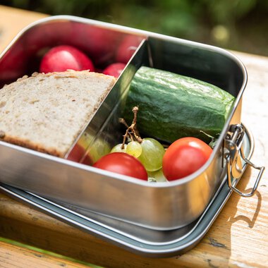 Eine Lunchbox mit frischem Brot, Gurke, Kirschtomaten, Trauben und Radieschen. Sie steht auf einem Holzuntergrund in einem natürlichen Umfeld. | © Kleinwalsertal Tourismus | Bastian Morell