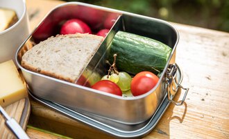 Eine Lunchbox mit frischem Brot, Gurke, Kirschtomaten, Trauben und Radieschen. Sie steht auf einem Holzuntergrund in einem natürlichen Umfeld. | © Kleinwalsertal Tourismus | Bastian Morell