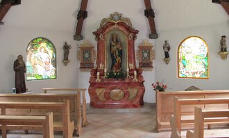 Eine kleine Kapelle mit Holzbankreihen und bunten Fenstern. Im Vordergrund steht ein schmuckvoller Altar mit einer Statue. | © Stefan Heim